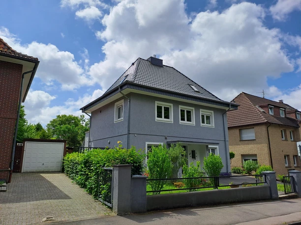 Ein modernes, graues Haus mit einem Satteldach und großen Fenstern. Grünpflanzen und ein offener Gartenbereich sind ebenfalls sichtbar.
