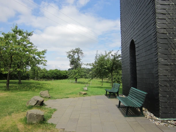 Eingang zur Windrather Kapelle, Velbert Zwei grüne Bänke stehen vor der schwarzen Fassade der Windrather Kapelle, umgeben von Bäumen.