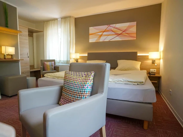 Hotel-Beckord-Altdeutscher Hof Modernes Hotelzimmer mit Doppelbett, Sitzbereich, Schreibtisch, Fenster und dezenter Wandkunst.