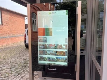 Infoterminal Bruchhausen-Vilsen Infoterminal mit Bildschirm vor einem Backsteingebäude, zeigt digitale Informationen und Fotos.
