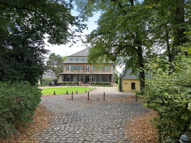 Schloss Eicherhof Das Bild zeigt ein herrschaftliches, von Bäumen umgebenes Herrenhaus mit einer gepflasterten Zufahrt.