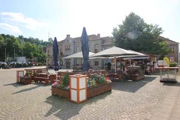 IMG_9063.JPG Ein öffentlicher Platz mit Sitzgelegenheiten, schattenspendenden Sonnenschirmen und umliegendem Grün.