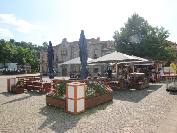 IMG_9063.JPG Ein öffentlicher Platz mit Sitzgelegenheiten, schattenspendenden Sonnenschirmen und umliegendem Grün.