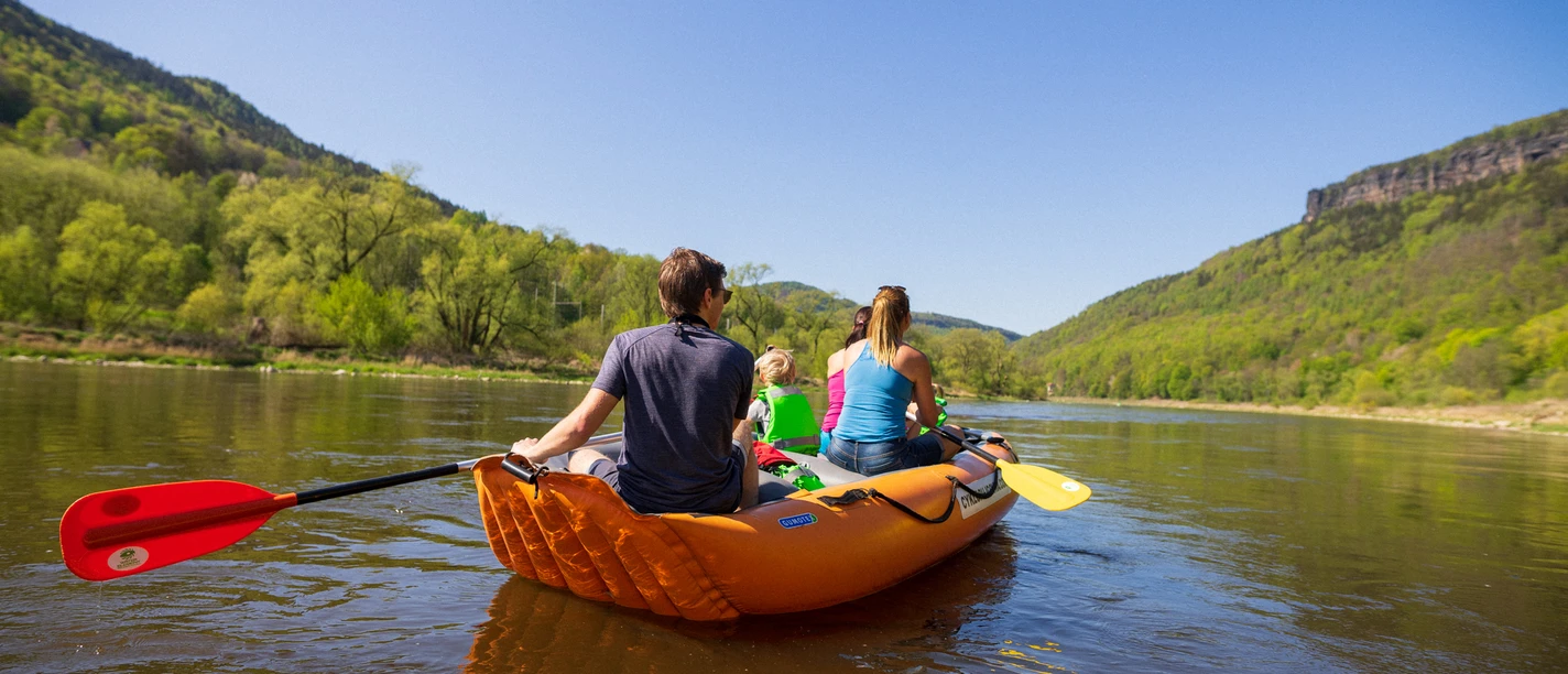 Fluss- und Bergwandertour - Packraft Packrafting auf einem ruhigen Fluss, umgeben von grünen Hügeln unter einem strahlend blauen Himmel.