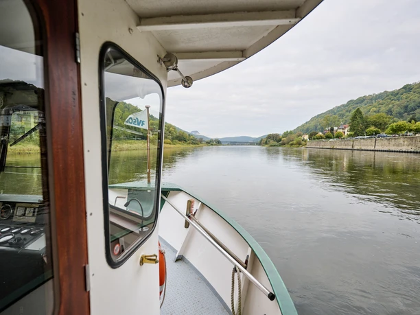 Uitzicht vanaf een veerboot op een kalme rivier, omringd door beboste heuvels en een stenen muur aan de oever; het is bewolkt weer.