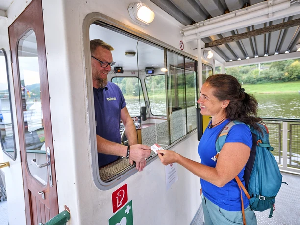 Im Gespräch mit dem Fährmann Eine Frau in blauem T-Shirt und Rucksack kauft ein Ticket bei einem Mann in einem Fährhäuschen. Beide lächeln, im Hintergrund ist Wasser zu sehen.