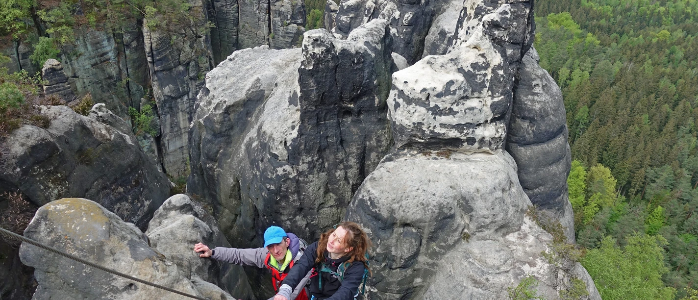 geführte Stiegentour Tourengeher erklimmen über Leitern steile Felsen in der Sächsischen Schweiz, umgeben von grünen Wäldern.