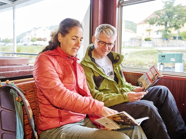 Een man en een vrouw zitten in een treincoupé, lezen brochures en dragen kleurrijke jassen; buiten is een landelijk landschap te zien.