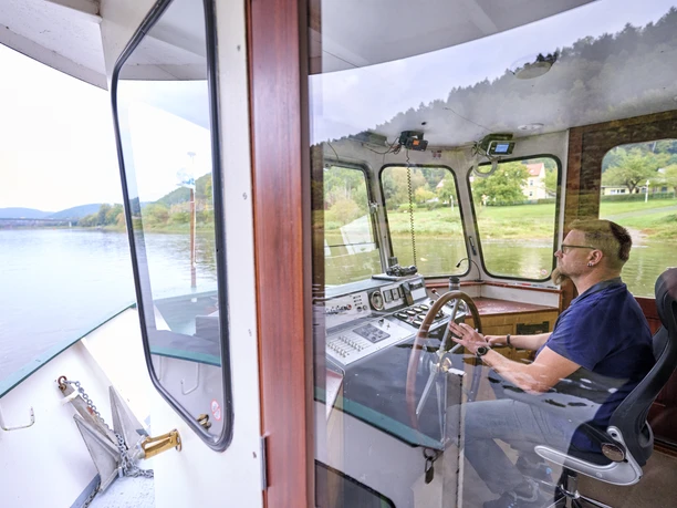 Een man gekleed in blauw bestuurt een boot op een rivier, omringd door groene landschappen en heuvels, zichtbaar door grote ramen.