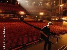 Ein Musiker mit Gitarre steht auf der Bühne eines leeren, prachtvoll beleuchteten Theatersaals.