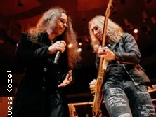 Zwei Musiker mit langen Haaren performen auf einer Bühne. Einer hält ein Mikrofon, der andere spielt Gitarre.