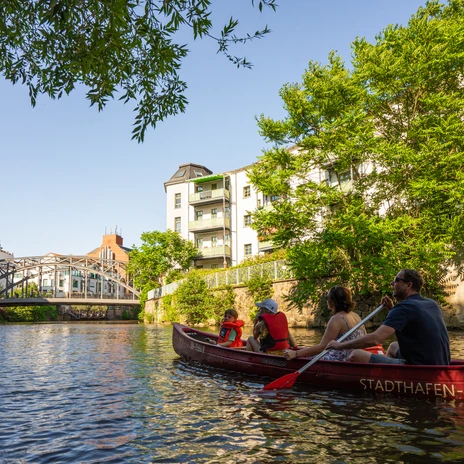 Rodzina z dwójką dzieci wiosłująca w czerwonym kajaku na Weiße Elster latem, w tle most Könneritz, Water City Leipzig