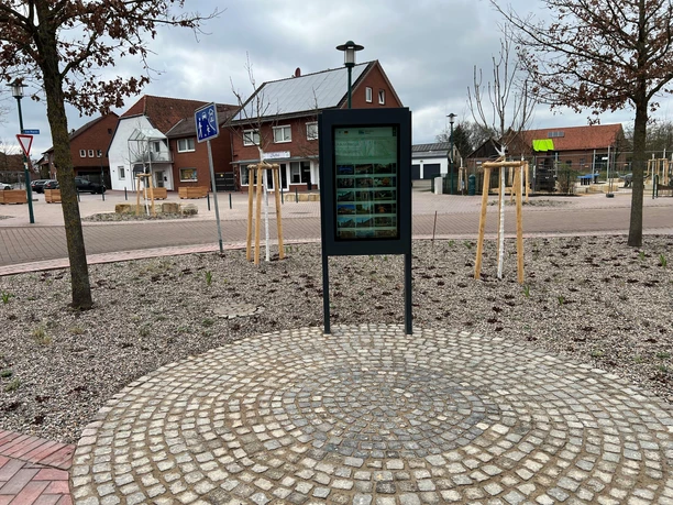 Information kiosk on a paved square in Loccum, surrounded by young trees and buildings in the background.