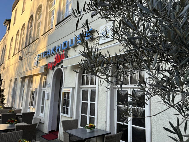 Herford-Steakhouse by Ilvi-Pro Herford GmbH.jpeg Steakhouse mit großer Fensterfront, modernen Olivenbäumen und Tischen auf dem gepflasterten Gehweg.