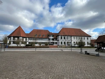 Fachwerkgebäude mit rotbraunen Dächern vor bewölktem Himmel, umgeben von Bäumen und gepflastertem Platz.