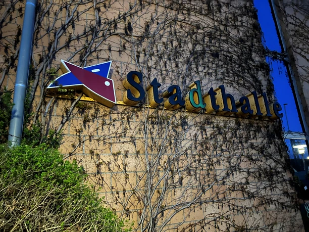 Ein bewachsener Außenbereich der Stadthalle Verden bei Dämmerung, mit markantem Schriftzug an der Fassade.