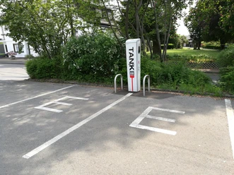 e-Tankstelle in Lindlar-Linde <p>Elektroladestation auf einem Parkplatz in Lindlar, umgeben von grünen Büschen und einem Gehweg.</p>