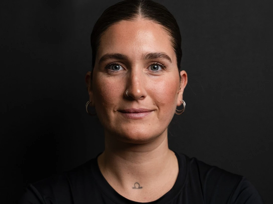 Pilates Rebecca Vahle Porträt einer Frau mit dunklem Haar und blauen Augen vor einem schwarzen Hintergrund.