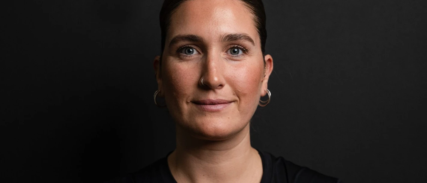 Pilates Rebecca Vahle Porträt einer Frau mit dunklem Haar und blauen Augen vor einem schwarzen Hintergrund.