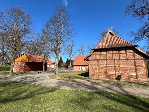 Ein traditioneller Fachwerkschafstall mit rotem Ziegeldach umgeben von kahlen Bäumen im ländlichen Umfeld.