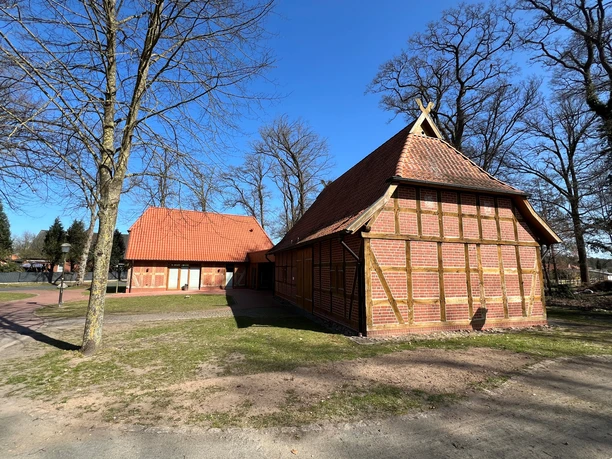 Fachwerkscheune mit roten Ziegeldächern umgeben von Bäumen vor klarem blauen Himmel in ländlicher Umgebung.