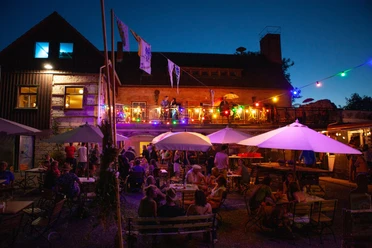 Heymannbaude A lively outdoor area at night with colorful fairy lights, lots of people at tables under white parasols and a stage in the background.