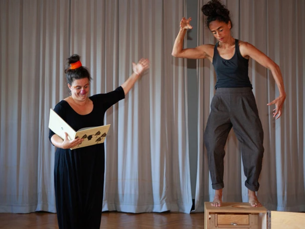 Aufführung Zaunkönig Festival Zwei Frauen in schwarzer Kleidung, eine liest aus einem Buch, die andere steht auf einem Holzkasten und gestikuliert, vor einem grauen Vorhang.