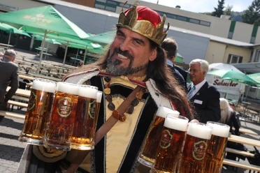 Gambrinus Mann in historischer Verkleidung trägt Bierkrugtabletts draußen auf einem Festgelände.