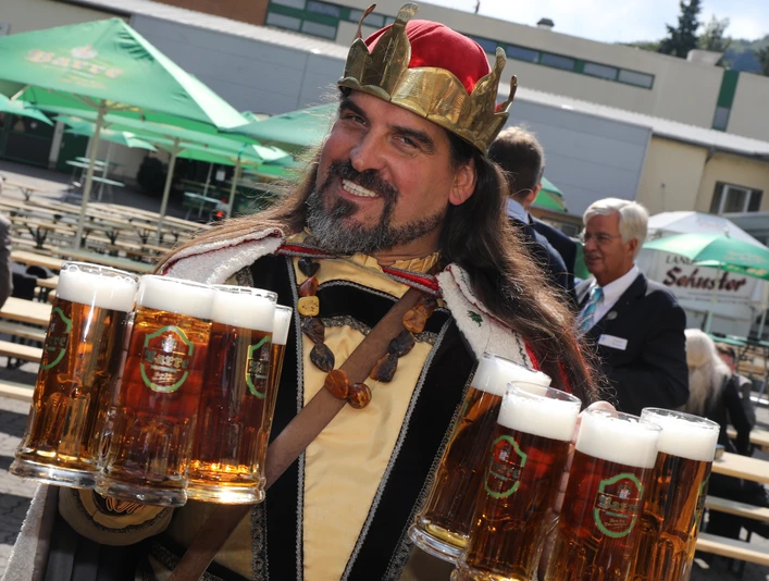 Gambrinus Mann in historischer Verkleidung trägt Bierkrugtabletts draußen auf einem Festgelände.