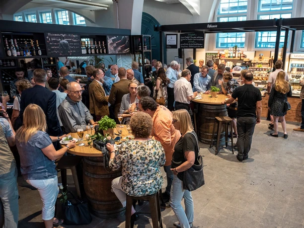 Geselligkeit in der Markthalle Herford_Pro Herford GmbH.jpg Eine geschäftige Markthalle mit Menschen, die an Holztischen gesellige Gespräche führen und genießen.