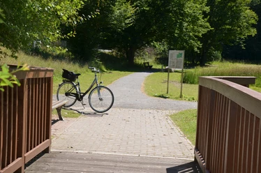 Symbolbild_Fahrrad_HD.JPG Geparktes Fahrrad an einer Holzbrücke. Im Grünen stehen Bäume und eine Infotafel.