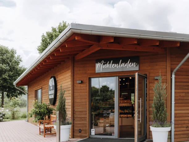 Holzgebäude mit Mühlenladen-Schild, offenem Eingang und Pflanzen, umgeben von Bäumen und Wolkenhimmel.