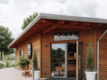 Ölmühle Solling Mülhenladen©Ölmühle Solling_Maike Wittreck.jpg Holzgebäude mit Mühlenladen-Schild, offenem Eingang und Pflanzen, umgeben von Bäumen und Wolkenhimmel.