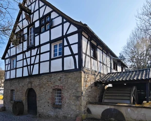 Rindersberger Mühle in Essen Fachwerkhaus aus Stein und Holz mit Wasserrad, umgeben von Bäumen, unter sonnigem Himmel.