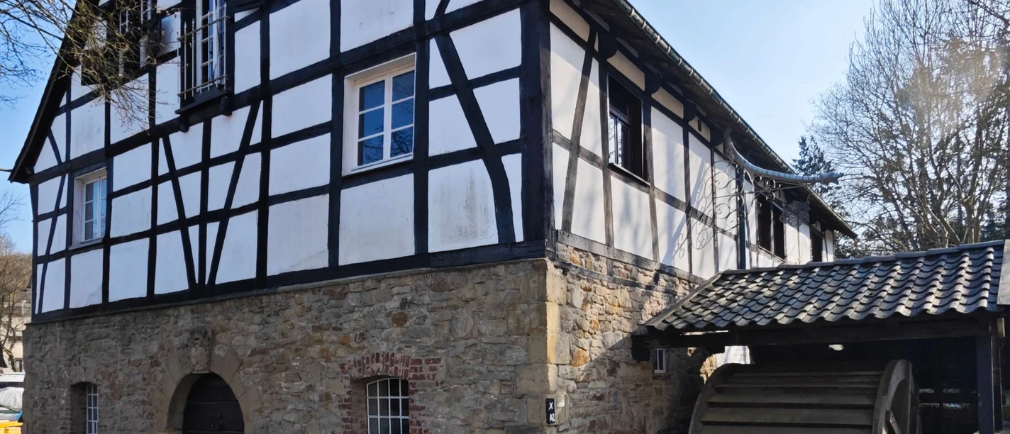 Rindersberger Mühle in Essen Fachwerkhaus aus Stein und Holz mit Wasserrad, umgeben von Bäumen, unter sonnigem Himmel.