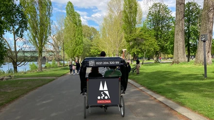 Rikolonia Picknick Rikscha Köln III.png Fahrradrikscha mit Fahrgästen in grünem Park, umgeben von Bäumen und Spaziergängern.