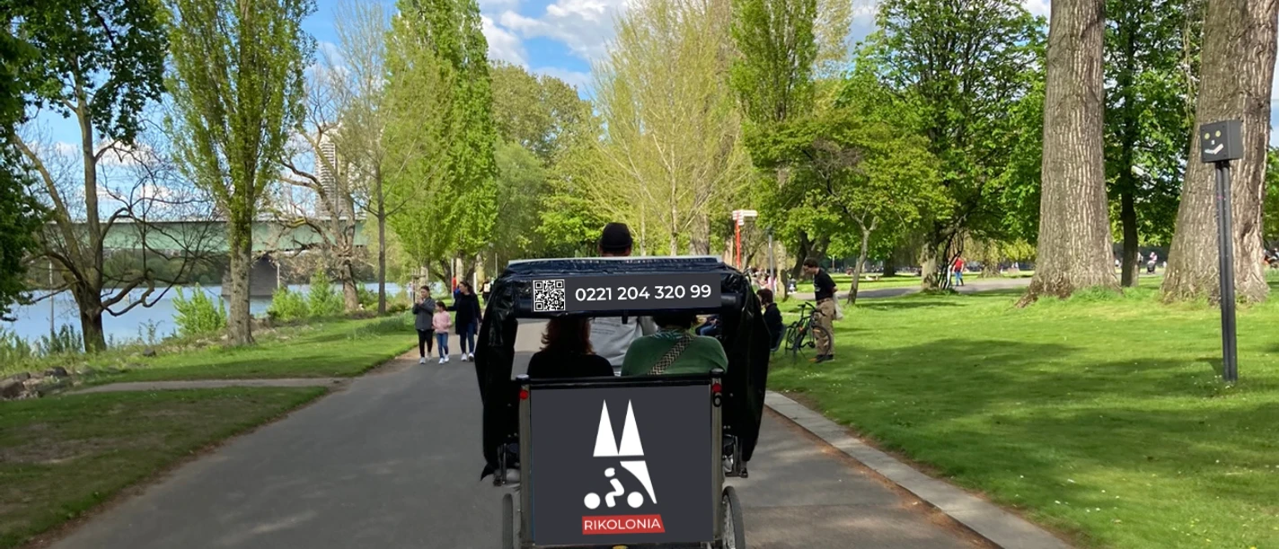 Rikolonia Picknick Rikscha Köln III.png Fahrradrikscha mit Fahrgästen in grünem Park, umgeben von Bäumen und Spaziergängern.