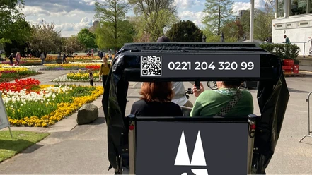 Rikolonia Picknick Rikscha Köln I.png Zwei Personen in einer Rikscha fahren durch einen Park mit bunten Blumenbeeten im Sonnenschein.