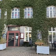 Herford-Gaststätte Bitter-Pro Herford GmbH (2).jpeg Eingewachsenes Gebäude mit großen Fenstern, Pflanzenfassade und Sitzgelegenheiten im Freien.