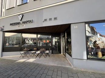 Herford-Pizzaria Napoli-Pro Herford GmbH (82).jpeg Eingang der Pizzeria Napoli mit bodentiefen Fenstern und Sitzgelegenheiten im Freien auf dem Gehweg.