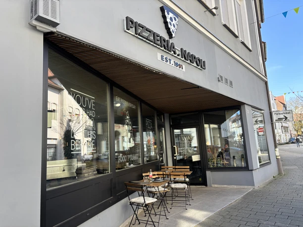 Herford-Pizzaria Napoli-Pro Herford GmbH (85).jpeg Pizzeria mit Außenterrasse, Holztischen und Stühlen, gelegen an einer Straßenecke, lädt zum Verweilen.