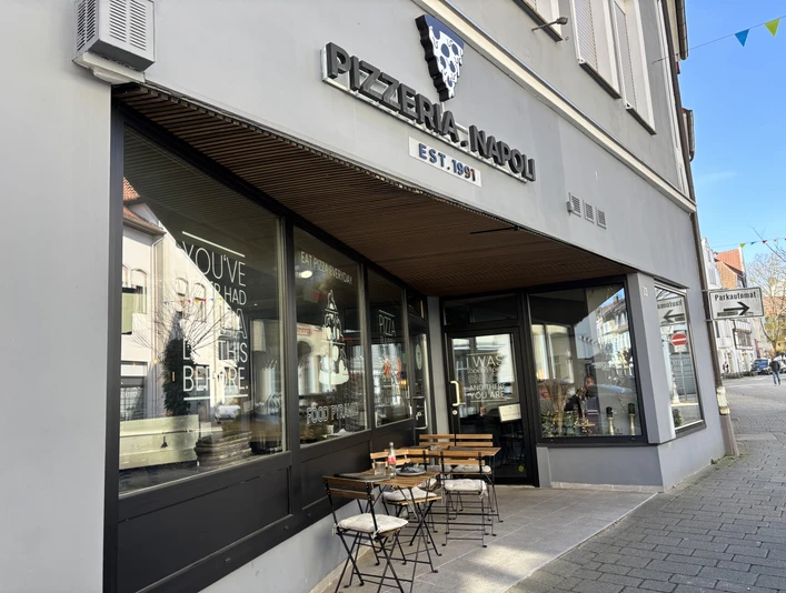 Herford-Pizzaria Napoli-Pro Herford GmbH (85).jpeg Pizzeria mit Außenterrasse, Holztischen und Stühlen, gelegen an einer Straßenecke, lädt zum Verweilen.