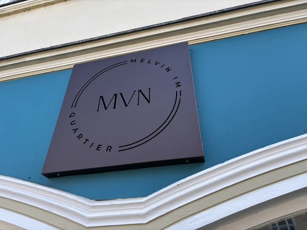Braunes Firmenschild von "Melvin im Quartier" auf türkisfarbenem Hintergrund, darauf das Logo MVN.