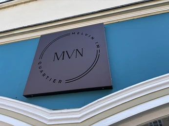 Herford-Melvin im Quatier-Pro Herford GmbH (68).jpeg Braunes Firmenschild von "Melvin im Quartier" auf türkisfarbenem Hintergrund, darauf das Logo MVN.