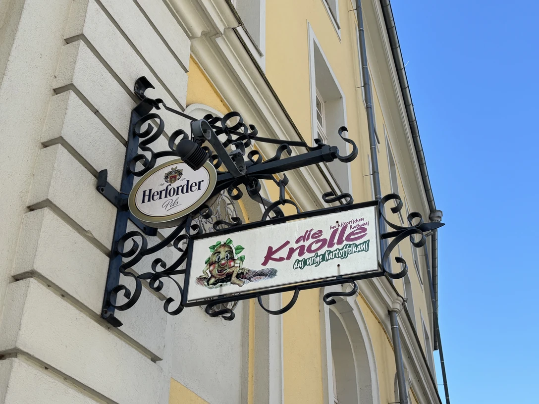 Herford-Die Knolle-Pro Herford GmbH (17).jpeg Metallschild an einer gelben Hausfassade mit dem Schriftzug "die Knolle" und einer gezeichneten Kartoffel.