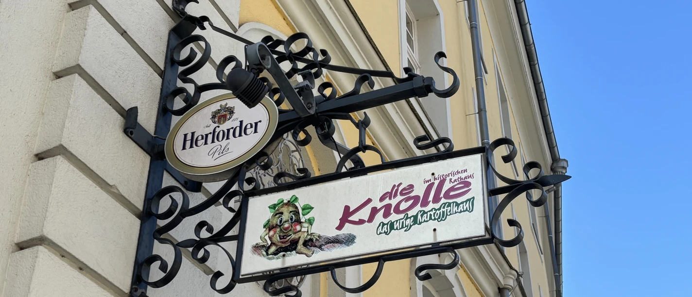 Herford-Die Knolle-Pro Herford GmbH (17).jpeg Metallschild an einer gelben Hausfassade mit dem Schriftzug "die Knolle" und einer gezeichneten Kartoffel.