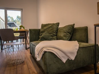 Bröcklingshof (8).jpg Grünes Sofa mit Kissen und Decke, daneben ein runder Couchtisch, im Hintergrund ein Esstisch vor Glasfront.