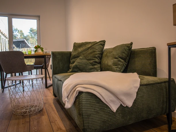 Bröcklingshof (8).jpg Grünes Sofa mit Kissen und Decke, daneben ein runder Couchtisch, im Hintergrund ein Esstisch vor Glasfront.