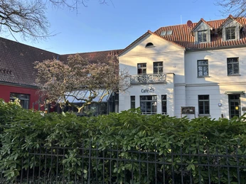 Herford-Café Hudl-Pro Herford GmbH (1).jpeg Historisches Gebäude mit weißer Fassade und quadratischen Fenstern, umgeben von grüner Hecke.