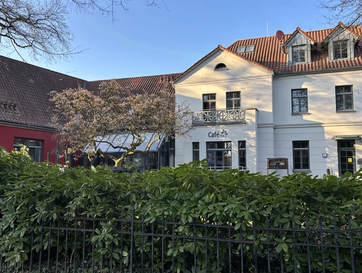 Herford-Café Hudl-Pro Herford GmbH (1).jpeg Historisches Gebäude mit weißer Fassade und quadratischen Fenstern, umgeben von grüner Hecke.